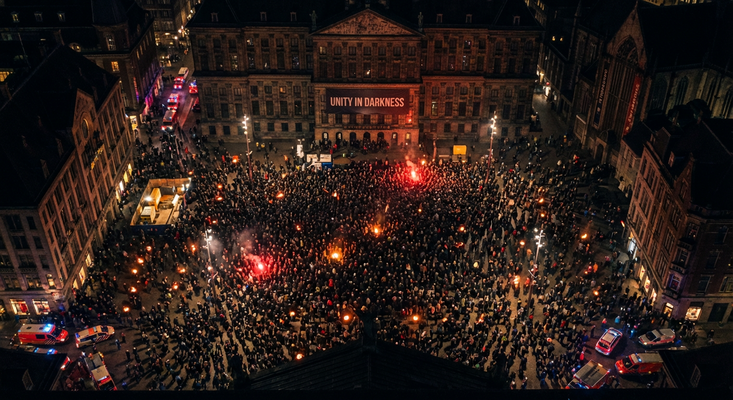 Aerial view of thousands of protesters gathered at night in a city square with burning red flares, beneath a 'UNITY IN DARKNESS' banner projected on the surrounding buildings